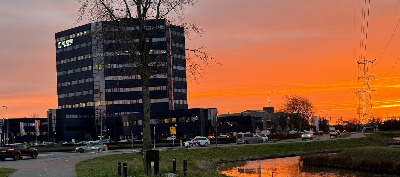 The OBV Group office building at dawn: a dark octagonal office tower silhouetted against an orange sunrise, with a canal and cycle path in the foreground.